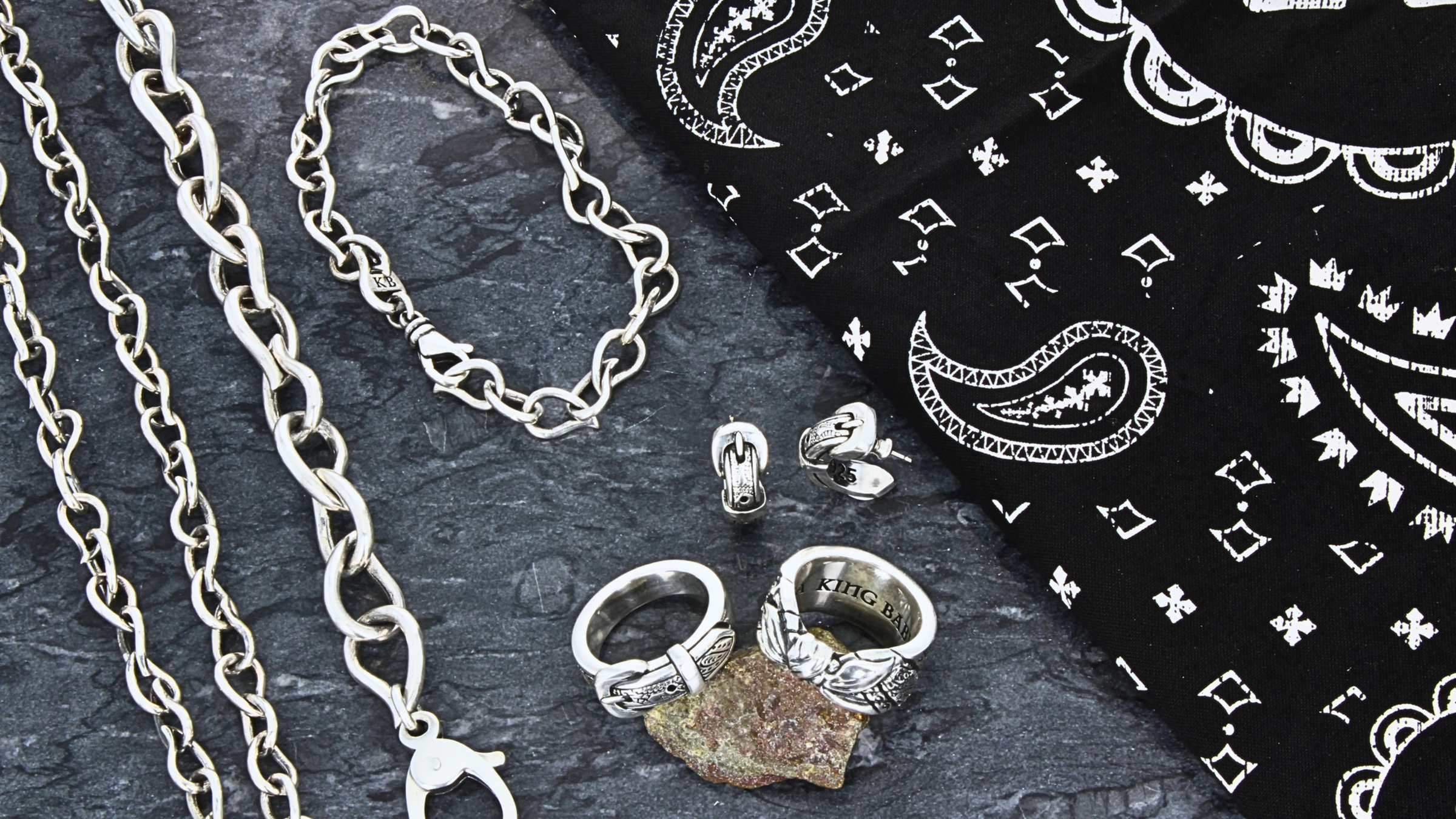 Silver jewelry including chains and rings on a dark surface with a bandana.